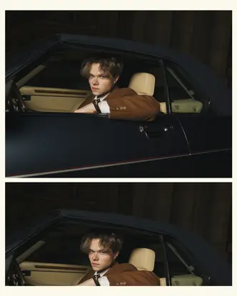 A person in a brown jacket sits in a car and looks through the open window.