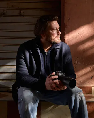 A man sits on a step with a camera in his hands, his face in the sunlight, in front of a closed shutter.