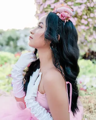 Woman in a pink dress and white gloves is sitting in a rose garden looking to the side.