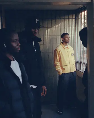 Five men in a shady hallway looking outside, the one in the middle wearing a yellow jacket.