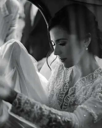 A bride sitting in a car holding her veil and a man standing next to the open car door.