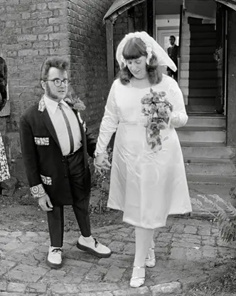 A wedding couple and a woman in a dress stand in front of a building.