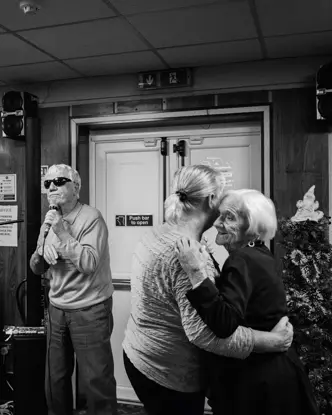 A man sings into a microphone while two older women dance and a man plays the keyboard in the background.