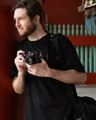 A photographer holds a camera while standing near traditional architecture, with two people in kimonos in the background.