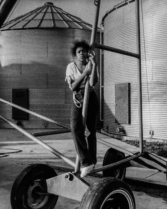 Young girl standing beside farm equipment and grain silos on a family farm in Arkansas, captured in black and white. A documentary photograph reflecting rural life, agricultural heritage, and generational connection.