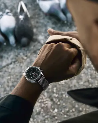 Jason Roman checks his Leica Watch while feeding pigeons in an urban public space, capturing a candid moment of everyday city life and human interaction with wildlife.
