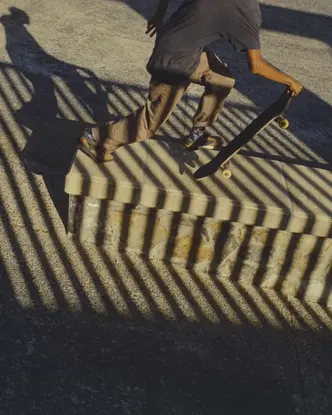 Skateboarder carrying a board through dramatic striped shadows at a skatepark, capturing movement, urban culture and graphic light.
