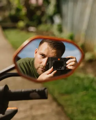 Photographer Philipp Gladsome reflected in a motorcycle mirror while taking a picture with a Leica M camera, surrounded by greenery in warm natural light.