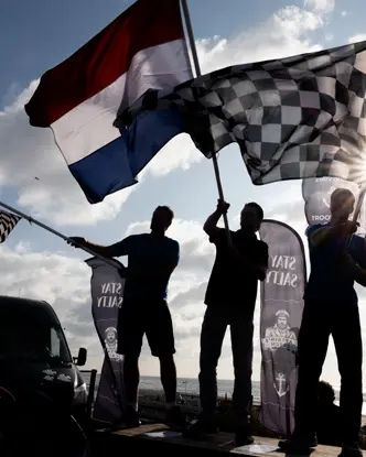 Silhouetted fans wave Dutch and chequered flags at a Formula 1 event in Zandvoort, with bright sunlight breaking through above the seaside circuit.