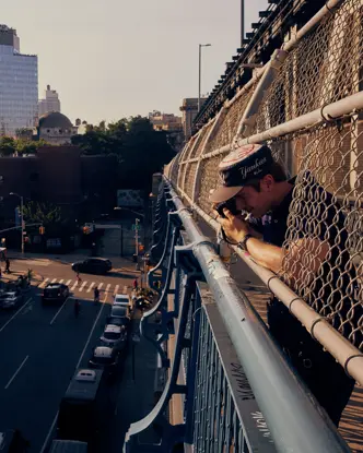 Paul Hepper leaning through the fence of a New York City bridge, taking a street photograph above traffic and urban buildings in warm evening light.