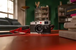 Leica camera with red strap on a table, blurred background with sofa and bookshelves.