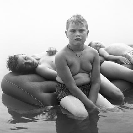 A boy sits in swimming trunks on an air mattress in the water.