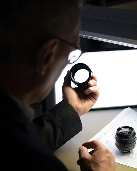 A man inspects a camera lens, technical drawings lie on the table.