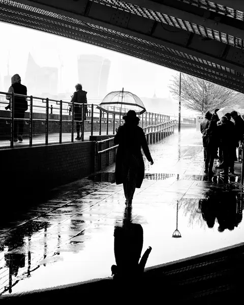 People walking under a bridge in the rain, silhouettes reflected in puddles.