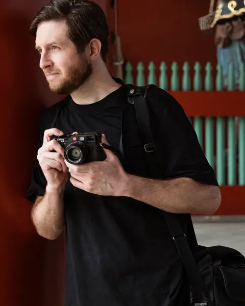 A photographer holds a camera while standing near traditional architecture, with two people in kimonos in the background.