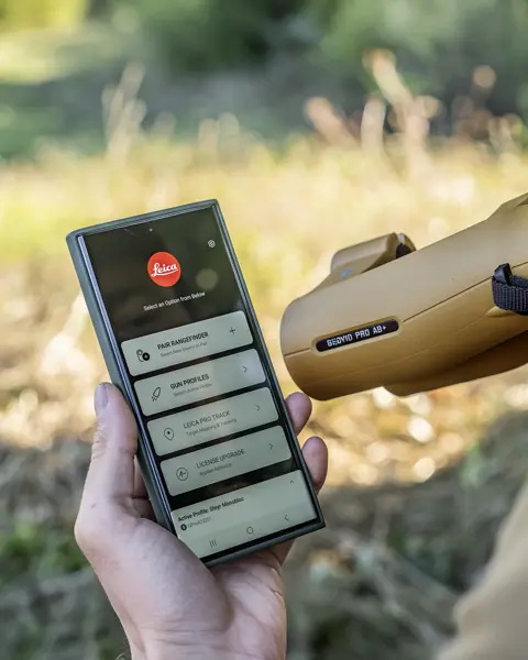 A mobile phone held by a hunter, opened with Leica Ballistics App and Leica Geovid Pro AB+ in the foreground, a meadow can be seen in the background.