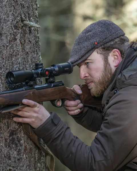 The Leica Fortis 6 1.8-12x42i is in focus while a hunter leans against a tree with his weapon and looks through the scope